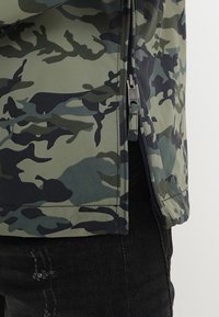 Giacca camouflage in tonalità di verde e nero, con zip laterale, texture liscia e vestibilità rilassata. Accentata con cuciture scure.