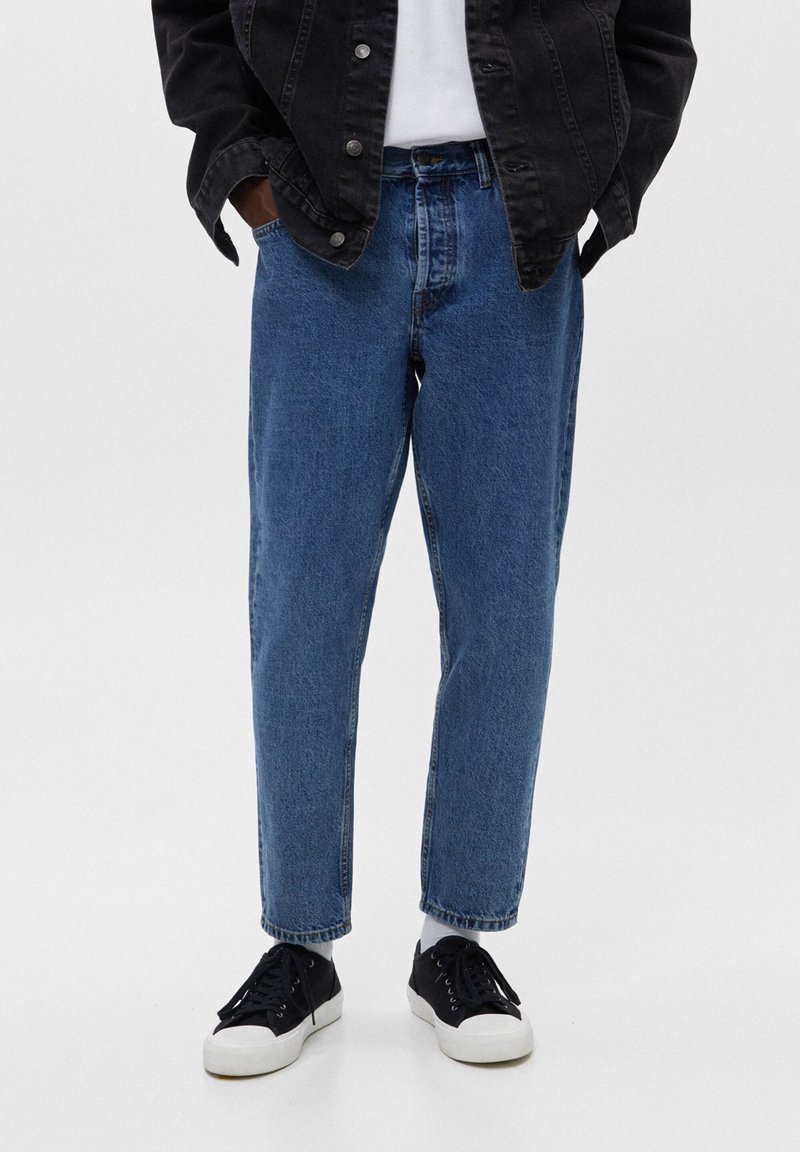 Vaqueros de mezclilla azul claro con un ajuste relajado, piernas rectas y un dobladillo sin rematar. Combinados con zapatillas negras y una chaqueta vaquera negra.