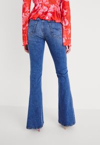 Mujer con jeans acampanados azules y una blusa de manga larga roja con flores, de pie contra un fondo blanco liso, vista desde atrás.