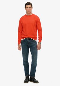 Czerwony sweatshirt z okrągłym dekoltem i ściągaczami na rękawach, noszony na białej koszulce, w połączeniu z niebieskimi dżinsami i czarnymi butami, stojący na białym tle.