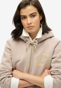 Beige ritshoodie met een trekkoord bij de nek, goud geborduurd logo en een witte binnenvoering. Zachte textuur, normale pasvorm.