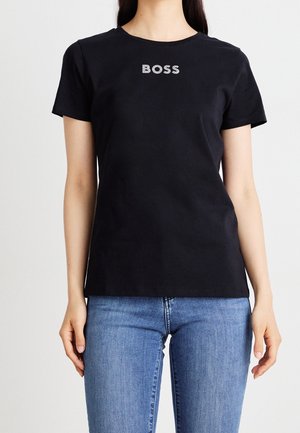Femme portant un T-shirt noir à manches courtes avec le texte « BOSS » sur la poitrine, associé à un jean en denim bleu clair, debout devant un fond uni.