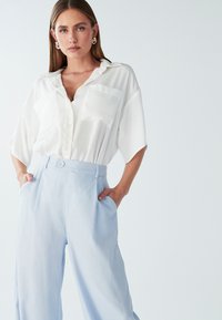 Bela kratka srajca s kratkimi rokavi in žepom, v kombinaciji z svetlo modrimi hlačami širokega kroja z višjim pasom, ki imajo sprednji gumb in stranske žepe.