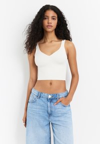 Fehér bordás crop top V-kivágással és széles pántokkal, világoskék magas derekú farmerrel párosítva. Simább textúra, minimalista stílus.