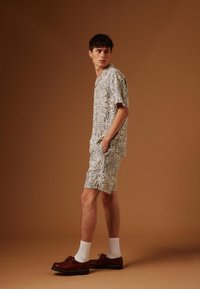 Jeune homme debout les mains dans les poches, portant une chemise à manches courtes à motifs et un short assorti, des chaussettes blanches et des chaussures en cuir marron.