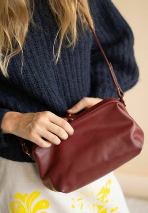 Piccola borsa in pelle borgogna con forma arrotondada, accessori dorati e una sola tracolla, tenuta da una persona che indossa un maglione di lana blu navy.