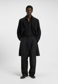 Cappotto di lana nera, camicia nera tessutata e pantaloni neri a pieghe si abbinano a scarpe nere. Il cappotto presenta un ampio colletto e spalle scese.