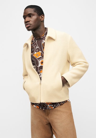 Άνδρας με μπεζ πλεκτό μπουφάν με φερμουάρ πάνω από καφέ και πορτοκαλί πουκάμισο με σχέδια, φοράει ανοιχτό καφέ παντελόνι, στέκεται με τα χέρια στις τσέπες μπροστά σε λευκό φόντο.