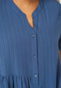 Blaue strukturierte Bluse mit V-Ausschnitt, kurzen Ärmeln und einem gestuften Design. Sie verfügt über fünf Knopfverschlüsse in passender Farbe.