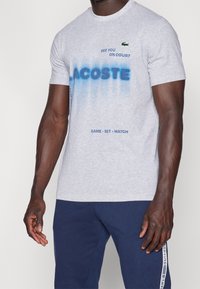 Grå bomulls t-shirt med blå spraymålad text, "LACOSTE," med grön krokodil-logga. Kombinerar med marinblå byxor som visar varumärkesaccenter.