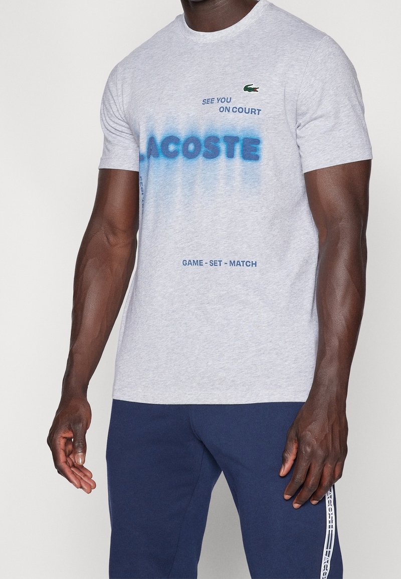 Grå bomulls t-shirt med blå spraymålad text, "LACOSTE," med grön krokodil-logga. Kombinerar med marinblå byxor som visar varumärkesaccenter.