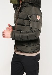 Grøn pufferjakke med aftagelig hætte, lynlås lukning, ribstrikkede manchetter og et logo-patch på venstre arm; glat, quiltet tekstur.