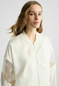 Weißer Bomberjacke mit transparenten Netzärmeln, Reißverschluss und geometrischem Paneeldesign mit transparenten Akzenten und subtiler Markenkennzeichnung.