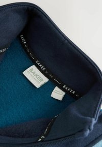 Cuello de sudadera marinera con forro suave en color teal, con etiquetas tejidas que dicen "BAKER by Ted Baker" y las instrucciones de cuidado. Textura suave, sin patrones.