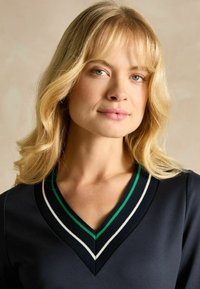 Marineblaue Bluse mit V-Ausschnitt, die einen grün-weißen gestreiften Saum aufweist. Glatter Stoff; lange, wellige Haare umrahmen das Gesicht. Natürlicher Makeup-Look.