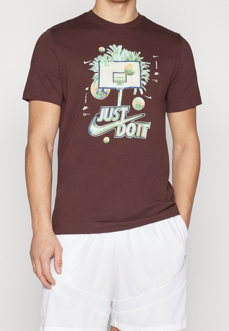 Homme portant un t-shirt Nike brun foncé avec un design d'arbre de palmier, un panier de basket-ball et le texte "Just Do It", assorti à un short de sport blanc.