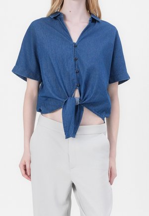 Vrouw draagt een blauw knoopoverhemd met korte mouwen, gestrikt in de taille, met hoge lichtbeige broek op een witte achtergrond.