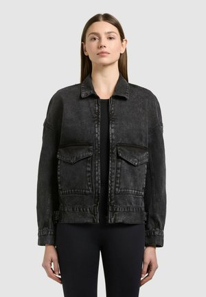 Femme portant une veste en denim noir délavé avec deux grandes poches à rabat à l'avant, debout devant un fond gris clair uni.