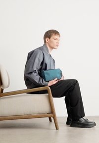 Teal veske med et enkelt design holdt i hånden, kombinert med en grå jakke og svarte bukser. Har glatt stoff og minimalt med hardware-detaler.