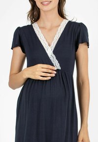 Granatowa sukienka kopertowa z krótkimi bufkami i dekoltem w kształcie litery V, zdobiona białą koronką wzdłuż dekoltu, wykonana z miękkiego materiału oraz mająca zwiewny fason.