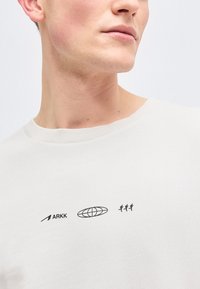 Κοντινό πλάνο ενός ατόμου που φοράει λευκό πουκάμισο με μαύρο λογότυπο ARKK, γραφικό γήινου και τρεις μικρές φιγούρες που τρέχουν στο στήθος.