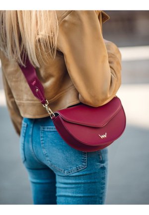 Bordó kožená kabelka přes rameno s křivkovým tvarem, doplněná zlatým logem. Nastavitelný popruh a hladká textura. Nositá přes hnědý kabát.