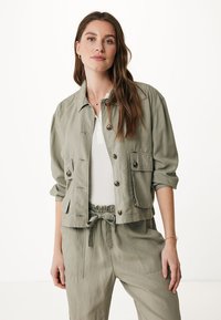 Donna con lunghi capelli castani che indossa una giacca leggera color oliva con bottoni su una maglietta bianca e pantaloni abbinati con cordoncino, contro uno sfondo semplice.