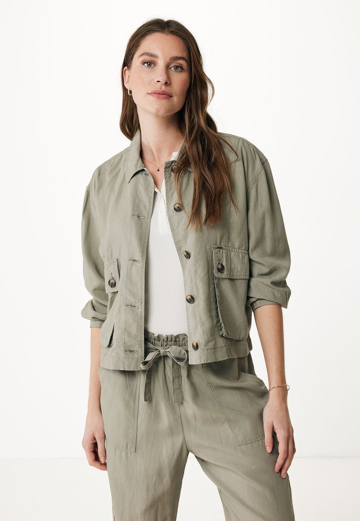 Donna con lunghi capelli castani che indossa una giacca leggera color oliva con bottoni su una maglietta bianca e pantaloni abbinati con cordoncino, contro uno sfondo semplice.