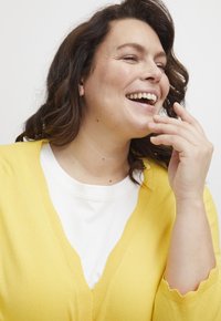Žlutý cardigan s výstřihem do V a vlnitým zakončením rukávů, vrstvený přes bílé tričko. Měkká struktura, lehký design.