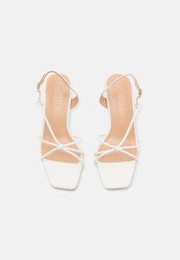 Escarpins blancs à talon ouvert avec des bouts carrés, dotés de fines brides et d'un design noué avec une semelle intérieure beige. Sangle de cheville réglable incluse.