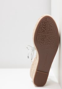 Sandały na koturnie z teksturowaną, naturalną górą i gładką brązową gumową podeszwą. Oferują białą koronkę oraz wytłoczone logo na podeszwie.