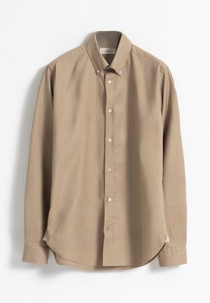 Camisa de manga larga de algodón beige. Presenta un cuello clásico, botones blancos y un dobladillo curvado con aberturas laterales para facilitar el movimiento.
