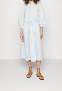 Abito azzurro chiaro a maniche lunghe con bottoni, dotato di una cintura in vita aderente e maniche a palloncino. Abbinato a sandali marroni con cinturini.