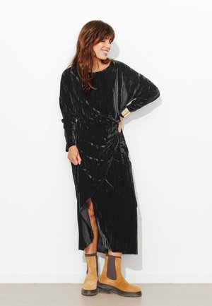 Robe portefeuille en velours noir à manches longues avec une fente latérale, associée à des bottines marron clair avec des panneaux élastiques et une semelle épaisse.