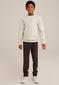 Krämfärgad sweatshirt med ribbstickade ärmar, tillsammans med mörkbruna texturerade byxor. Modellen har vita sneakers och står framför en neutral bakgrund.