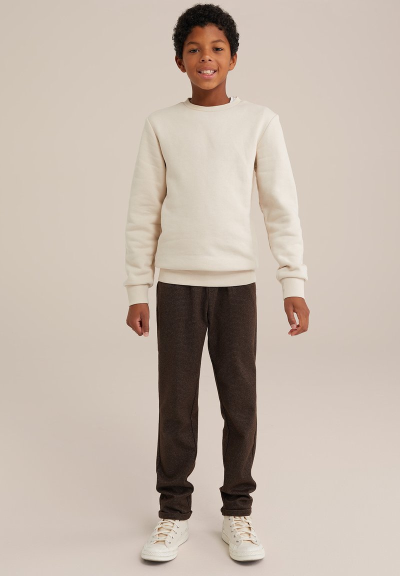 Krämfärgad sweatshirt med ribbstickade ärmar, tillsammans med mörkbruna texturerade byxor. Modellen har vita sneakers och står framför en neutral bakgrund.
