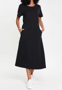 Robe midi noire à manches courtes avec une taille cintrée et des poches latérales. Fabriquée en tissu doux à la texture lisse. Associée à des baskets blanches.