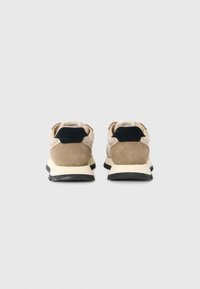 Ett par beige och svarta sneakers sedda bakifrån, med mockadetaljer, tjocka vita sulor och svarta hälpatcher.