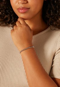 Metalen armband met kleine ronde kralen, zilverkleurig, gedragen om een pols. Close-up van een hand die bij de kin rust, met de nadruk op het ontwerp van de armband.