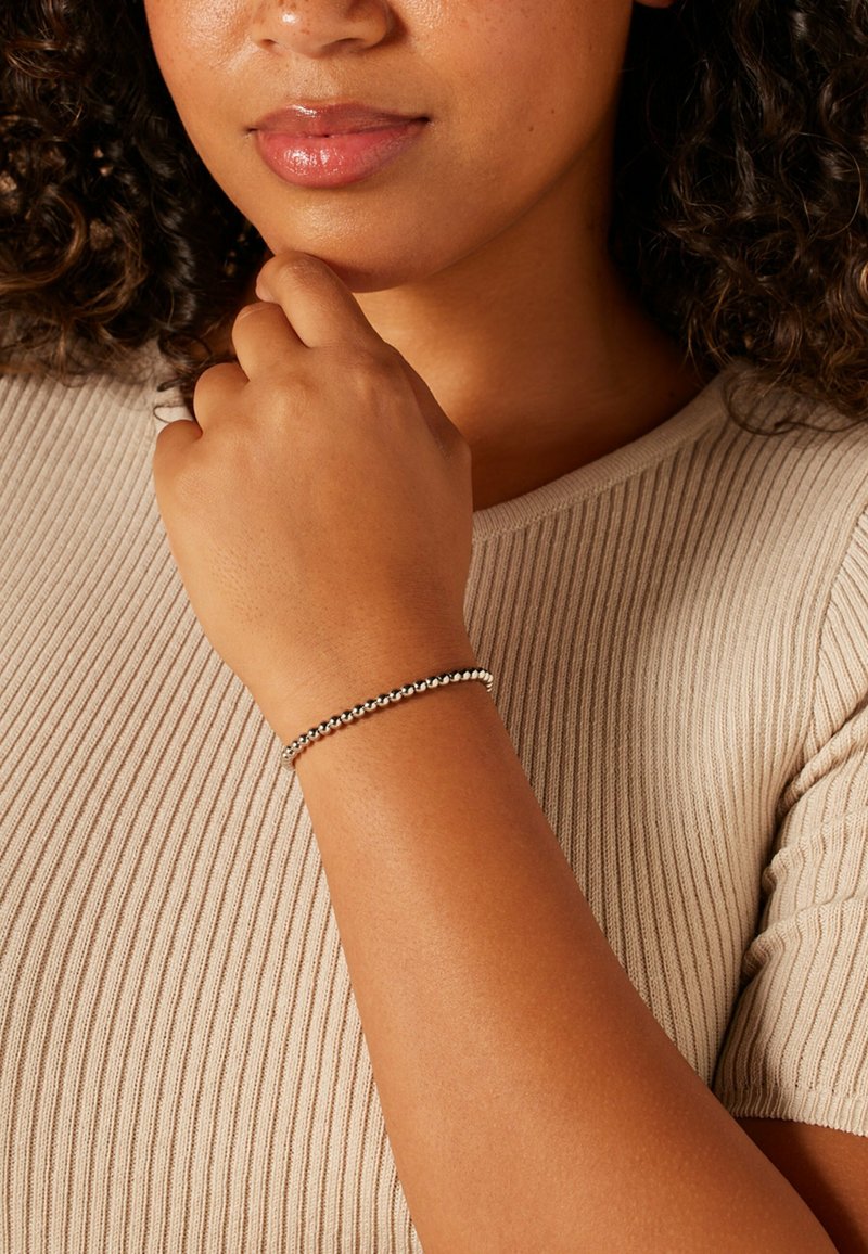 Metalen armband met kleine ronde kralen, zilverkleurig, gedragen om een pols. Close-up van een hand die bij de kin rust, met de nadruk op het ontwerp van de armband.