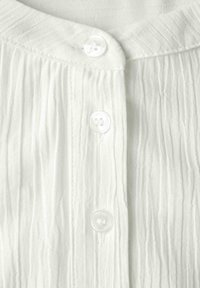 Chemise blanche avec un design texturé à plis, ornée de quatre boutons blancs à l'avant et d'un col arrondi.