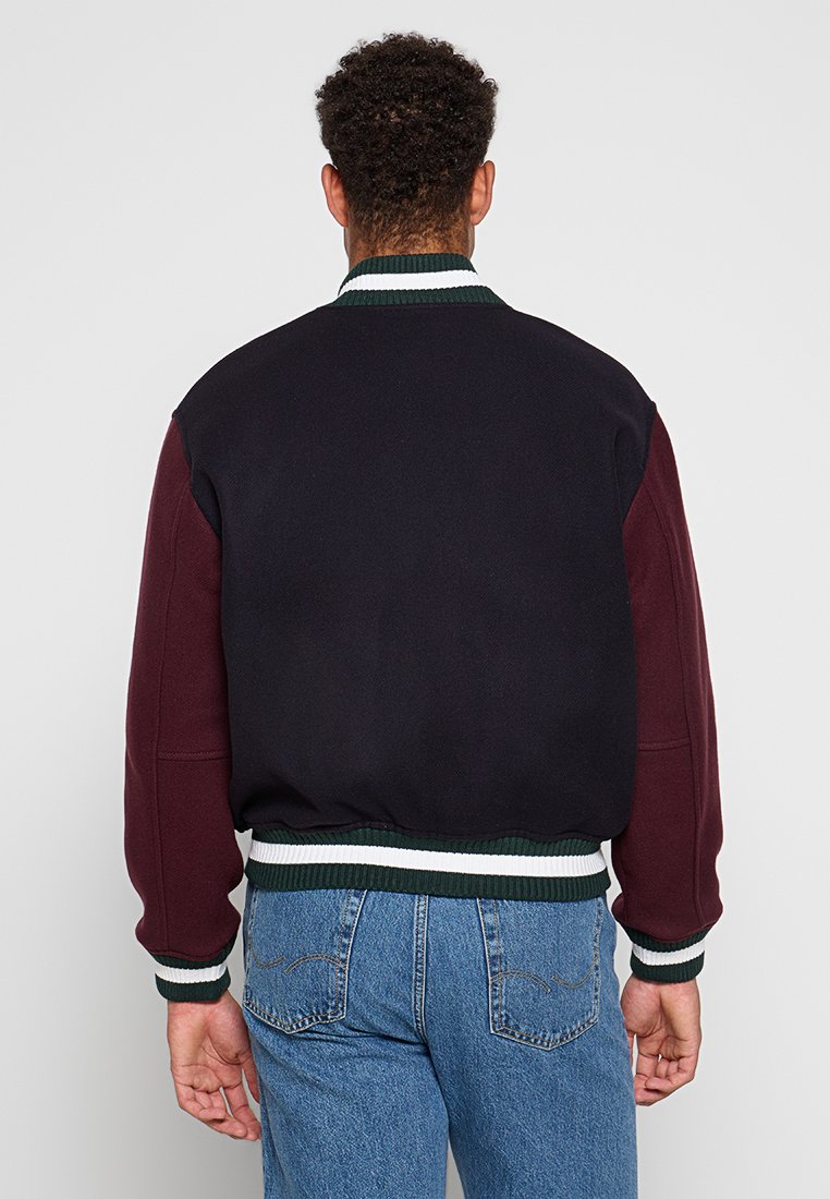 Veste bomber pour homme avec un design color-block en bleu marine et bordeaux, dotée de poignets et d'une taille côtelés verts et blancs, en tissu doux.