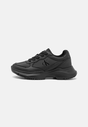 Baskets de sport noires à lacets sur le devant, semelle épaisse et logo « cK » sur le côté, sur un fond clair uni.
