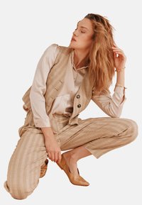 Beige geruite vest, bijpassende broek en licht satijnen blouse. Gelaagde bronzen instappers. Model poseert op de knieën met losjes gestyled haar.