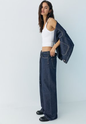 Nő fehér ujjatlan felsőben, széles szárú sötét kék farmernadrágban, egyik vállán egy hozzá illő farmerdzsekit tartva, semleges háttér előtt.