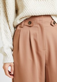 Béžový pletený svetr přes světle hnědé kalhoty s plisováním, bočními kapsami a detailními knoflíky. Hladká textura a přiléhavý střih.