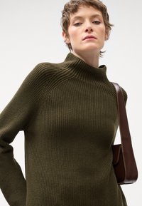 Maglione a coste verde oliva con colletto alto e maniche lunghe; presenta una silhouette aderente ed è indossato con una borsa a tracolla marrone.