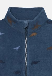 Giacca in pile con zip di colore blu scuro e colletto alto. Presenta motivi di dinosauri multicolori in marrone e azzurro chiaro. Tessuto morbido.
