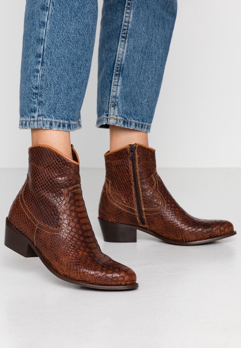 Botas de tobillo marrones con textura y cremalleras laterales, llevadas con jeans azules remangados sobre un suelo blanco.