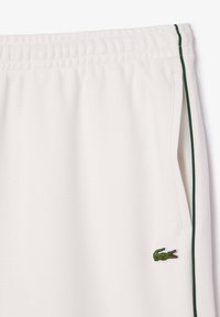 Pantalon de jogging blanc avec une finition texturée, comportant un logo crocodile vert et un liseré vertical vert le long des poches latérales.